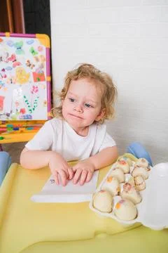 Child decorating Easter eggs with colorful stickers on yellow table at home. Stock Photos
