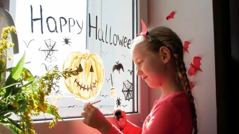 Child in devil horns painting pumpkin on window preparing celebrate Halloween Stock Footage 161012265