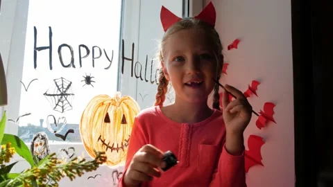 Child in devil horns painting pumpkin on window preparing celebrate Halloween Stock Footage 161013071