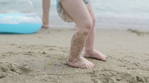 Child in diaper plays on sandy beach, digging with orange shovel, then stands Stockbeeldmateriaal 340900882