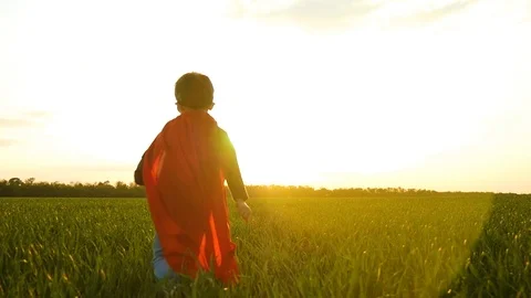 A child dressed as a superhero runs across the green grass towards the sunset Stock Footage