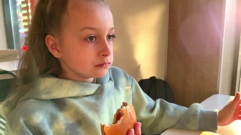 The child eats a big burger in a fast food restaurant, unhealthy food, the girl Stock Footage 241080322
