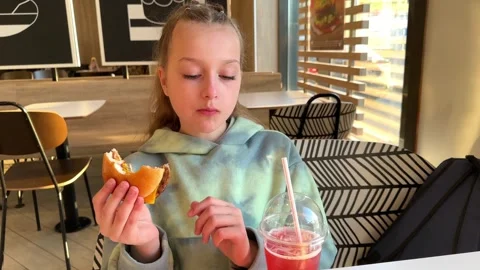 The child eats a big burger in a fast food restaurant, unhealthy food, the girl Stock Footage 241080379