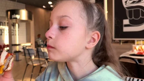 The child eats a big burger in a fast food restaurant, unhealthy food, the girl Stock Footage 241080396