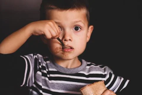 The child eats ice cream with a spoon. The child eats on a dark background. B Stock Photos