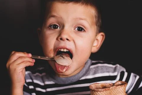 The child eats ice cream with a spoon. The child eats on a dark background. B Stock Photos
