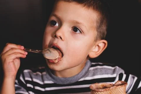 The child eats ice cream with a spoon. The child eats on a dark background. B Stock Photos