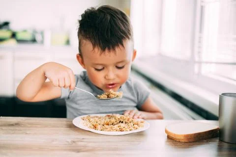 The child eats pilaf with carrots in the kitchen during the day Stock Photos