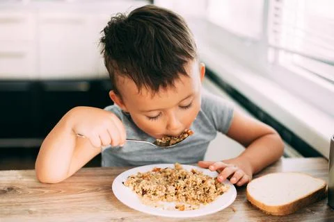 The child eats pilaf with carrots in the kitchen during the day Stock Photos