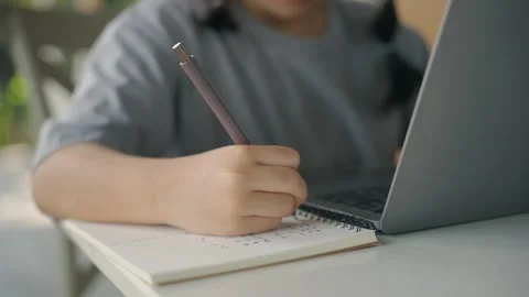 Child Engaged in Homework, Writing in Notebook, Using a Laptop, Focused on .. Stock Footage 333912223