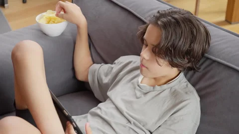 Child engrossed in a digital tablet while eating snacks lying on a cozy couch Stock Footage 315560399