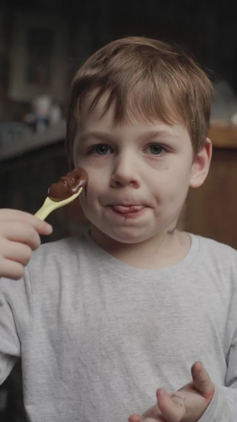 Child enjoys chocolate spread while licking spoon in cozy kitchen setting Stock Footage 297076889