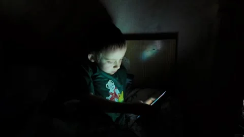 Child focused on smartphone in a dark room while sitting on a bed in the evening Video stock 338437846