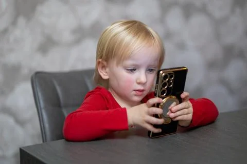 Child focused on smartphone while sitting at a table in a modern room Stock Photos