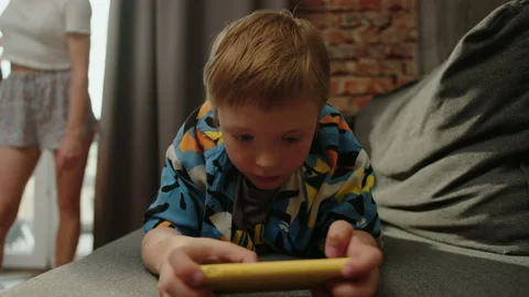 A child gamer lies on the couch while a young mother cleans the apartment Stock Footage 200989786