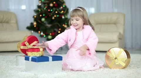Child gets a gift from the box. She sits beside the Christmas tree. Video stock 8975184