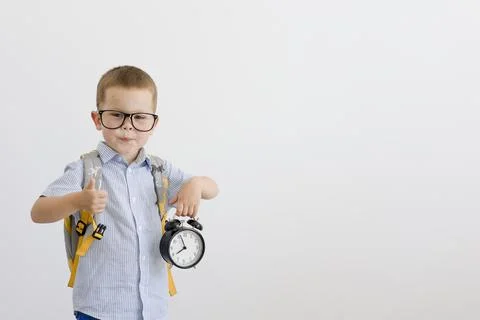 A child in glasses with a backpack on his back holds an alarm clock. Stock Photos