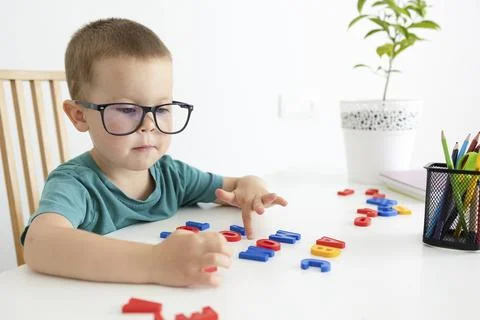 A child with glasses at a white table puts glory out of letters. Stock Photos