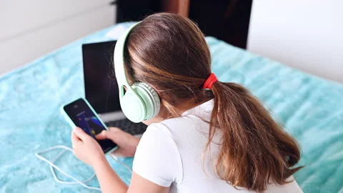 A child with headphones playing a computer game on the phone. A little girl lies 스톡 동영상 142104454