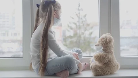 A child with her teddy bear both in masks stay by the window in the room Stock Footage 127579358