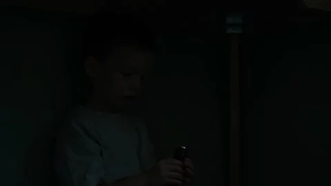 Child hiding beneath the table at night holds a torch and lights his own face Vídeo Stock 346305708