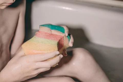 Child holding a multicolored sponge while sitting in a bathtub filled with .. 库存照片