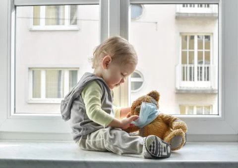 Child in home quarantine playing at the window with his sick teddy bear weari Stock Photos