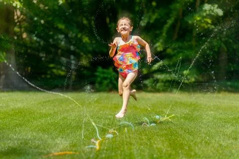 Child jumping and playing through a sprinkler in a backyard on a sunny day Stock Photos