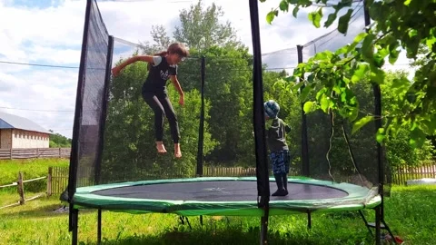 Child jumps on a trampoline in backyard with trees and fence on a sunny day Stock Footage 327319891