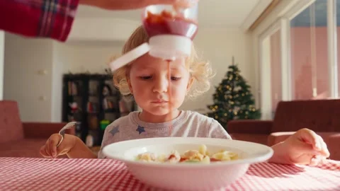 The child in the kitchen at the table eating macaroni with ketchup. Mother adds Stock Footage 228719711