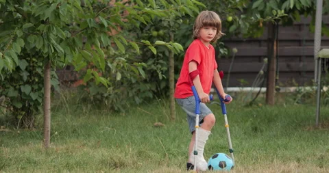 Child with leg in cast stands on crutches, playing with soccer ball in garden Stock Footage 298857341