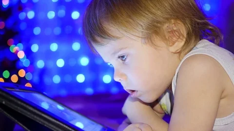 The child looks to the tablet lying on bed. In the background, lights and Video stock 70785993