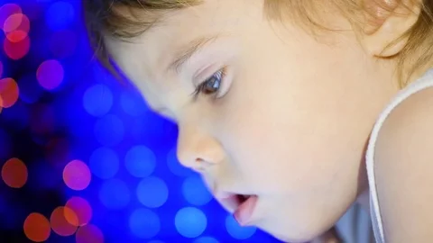 The child looks to the tablet lying on bed. In the background, lights and Видео 70787745