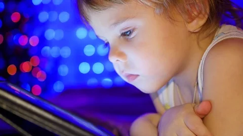 The child looks to the tablet lying on bed. In the background, lights and 스톡 동영상 70814811