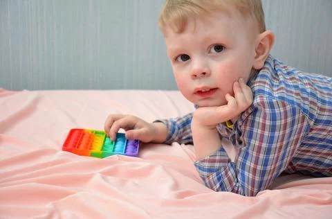 The child is lying on the bed with a POP IT toy. Stock Photos