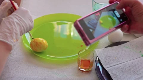 Child painting yellow Easter egg with brush on green plate while person takes Stock Footage 299403507