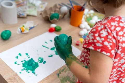 Child paints Easter eggs with green paint and makes handprints on paper at ho Stock Photos