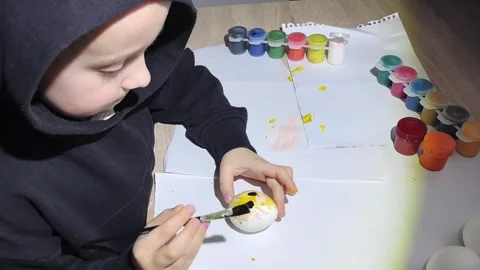 Child paints an egg with bright colors at a table with paint cups in a well-lit Stock Footage 328441311