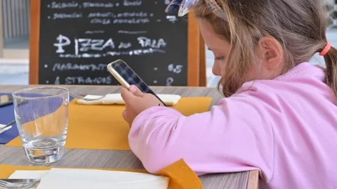 A child with a phone at a cafe, restaurant table. Little Girl Stock Footage 131697256