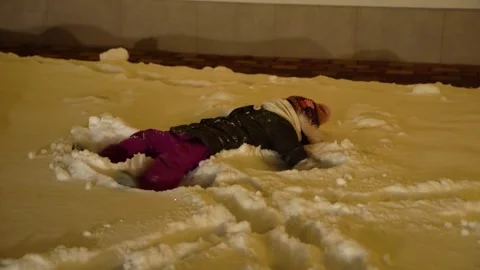 Child playing in fresh snow creating a snow angel with arms and legs moving at Stock Footage 328986946