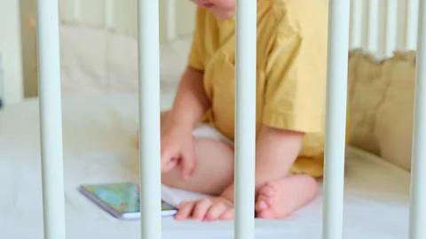 Child playing with mobile phone inside a white crib. Little boy using digit.. Stock Photos