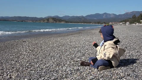 Child plays alone sitting on the pebble beach in winter Stock Footage 124698914