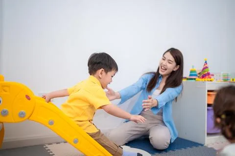 Child plays on slide while teacher welcomes him with open arms in classroom.. Stock Photos