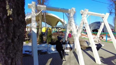 Child plays on swings at park on a sunny day with other kids and families around Stock Footage 332066853