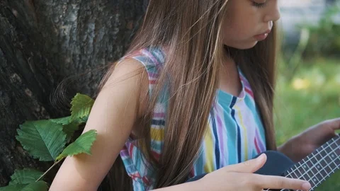 Child plays on ukulele sitting under a tree in a summer park. Stock Footage 115434945