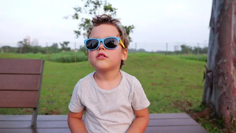 Child posing and make faces, wearing sunglasses, sits on bench in park Stock Footage 303624455