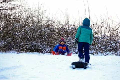 Child pulling black plastic sled in snow with another person sitting on red sled Stock Photos