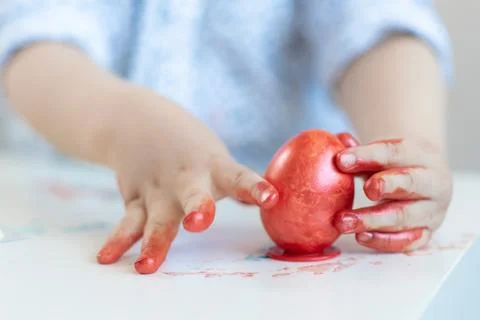 A child puts a red Easter egg on a stand with his hands stained with paint on Stock Photos