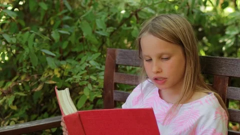 Child reading book aloud while sitting on a wooden bench in a park Stock Footage 304149286