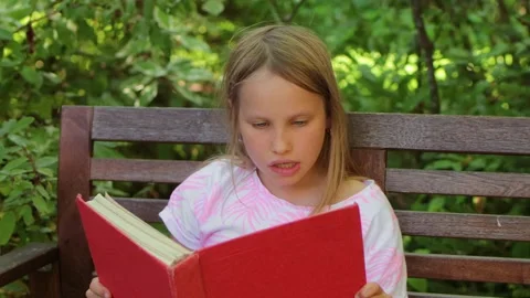 Child reading book aloud while sitting on a wooden bench in a park Stock Footage 304149287